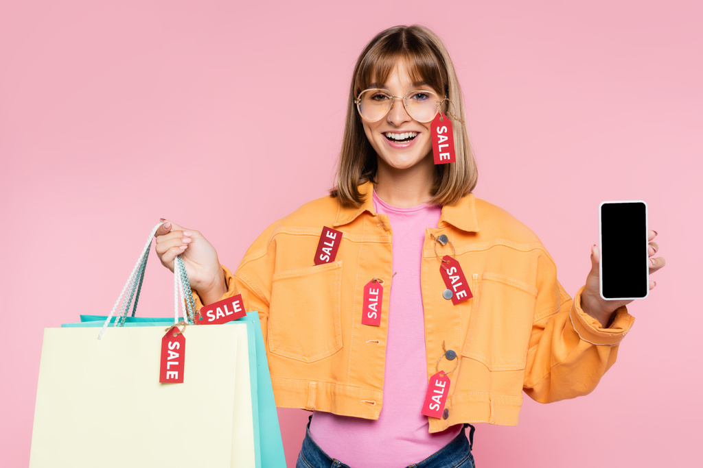 Young woman holding shopping bags with price tags and sale lettering and smartphone with blank screen on pink background - Photo, Image