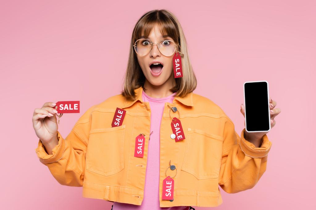 Έκπληκτος γυναίκα κρατώντας ετικέτα τιμή με λέξη πώληση και smartphone με λευκή οθόνη σε ροζ φόντο - Φωτογραφία, εικόνα