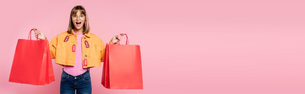 Imagem horizontal da mulher excitada com etiquetas de preço na jaqueta e óculos de sol olhando para a câmera enquanto segurava sacos de compras vermelhos no fundo rosa - Foto, Imagem