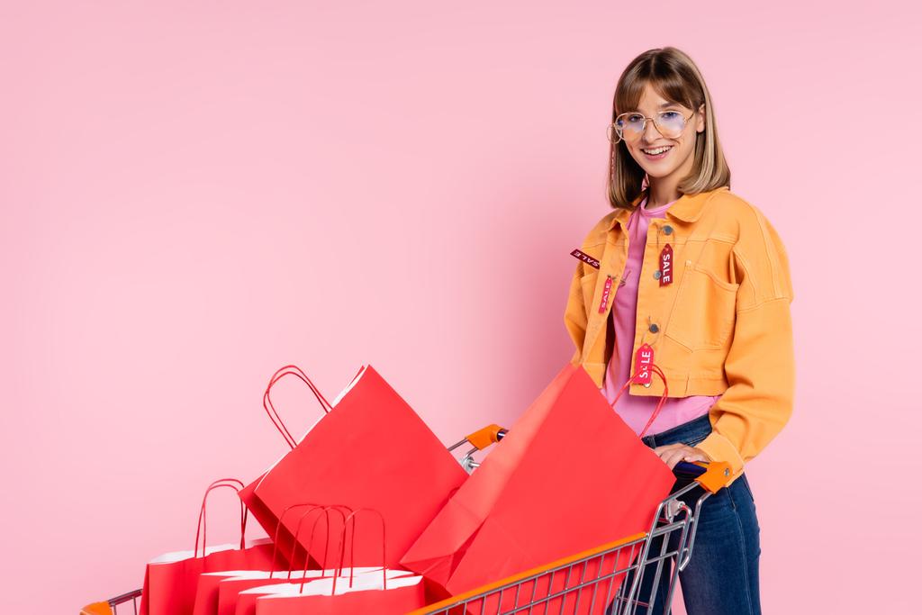 Young woman in sunglasses with price tags on jacket standing near cart with shopping bags on pink background - Photo, Image