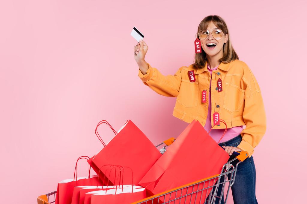 Mujer emocionada con etiquetas de precio en la chaqueta que sostiene la tarjeta de crédito cerca de las bolsas de la compra en el carro sobre fondo rosa - Foto, imagen