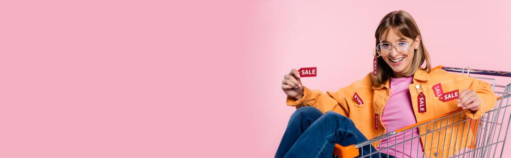 Πανοραμική έννοια της γυναίκας που δείχνει τις ετικέτες των τιμών με λέξη πώληση, ενώ κάθεται στο καλάθι σε ροζ φόντο - Φωτογραφία, εικόνα