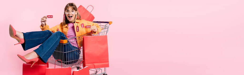Coltivazione orizzontale di donna sorpresa con cartellini dei prezzi guardando la fotocamera nel carrello con borse della spesa su sfondo rosa - Foto, immagini