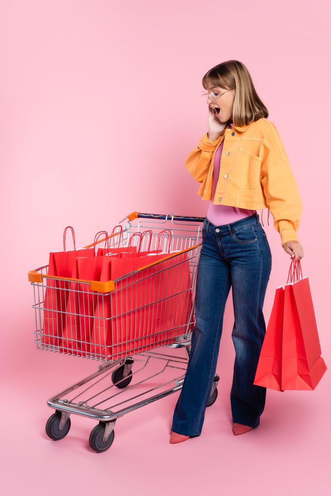 Verrast vrouw met de hand in de buurt wang kijken naar winkeltassen in winkelwagentje op roze achtergrond - Foto, afbeelding