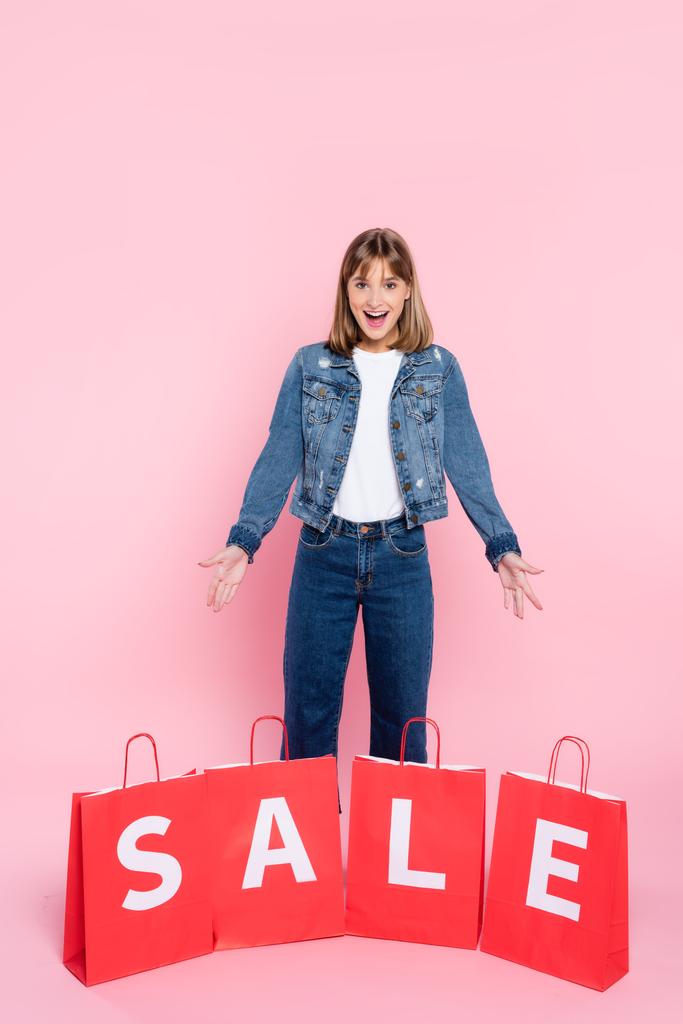 Young woman pointing with hands at shopping bags with sale lettering on pink background - Photo, Image
