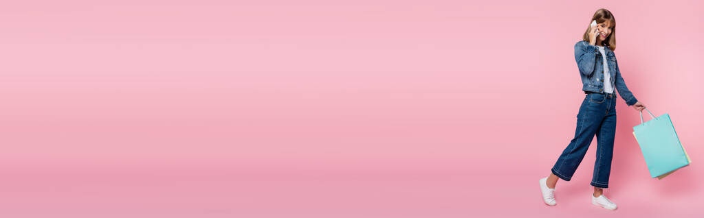 Panoraama laukaus nainen denim takki puhuu älypuhelimeen ja tilalla ostoskassit vaaleanpunainen tausta - Valokuva, kuva