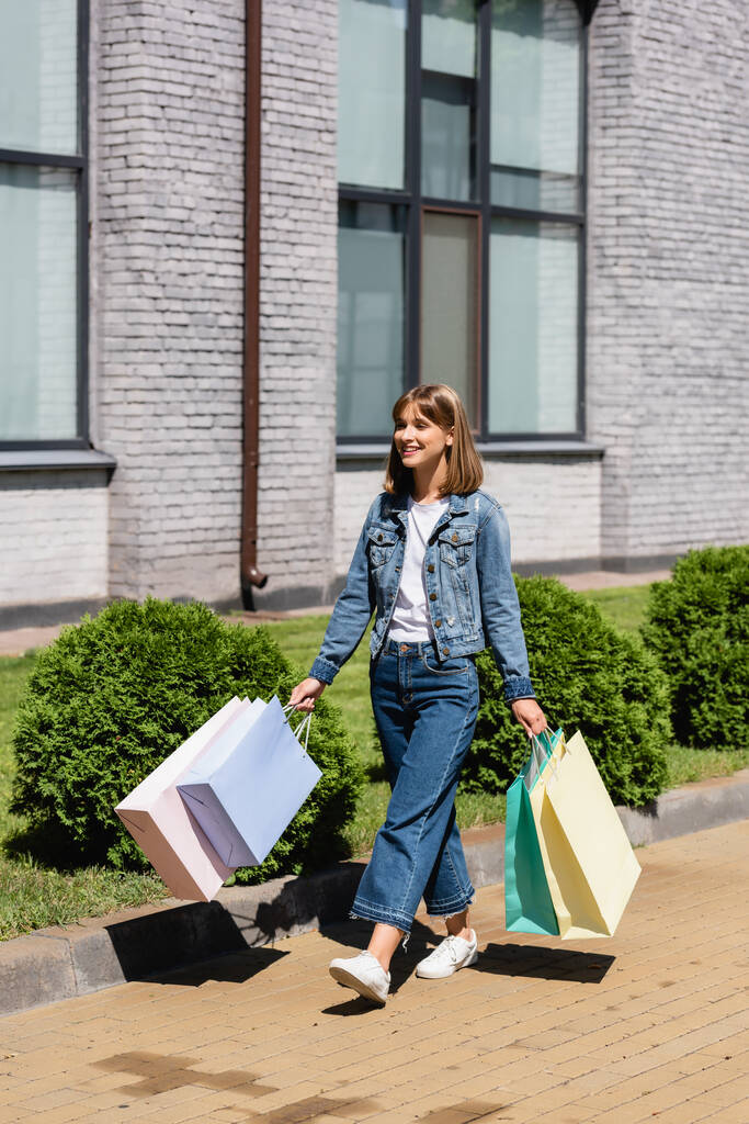 Jovem mulher de jaqueta jeans segurando sacos de compras enquanto caminhava na rua urbana  - Foto, Imagem