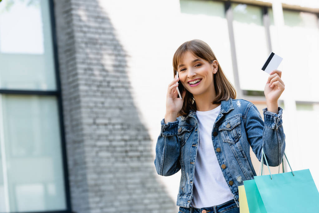 Junge Frau zeigt Kreditkarte, während sie Einkaufstüten hält und auf der Straße mit dem Smartphone spricht  - Foto, Bild