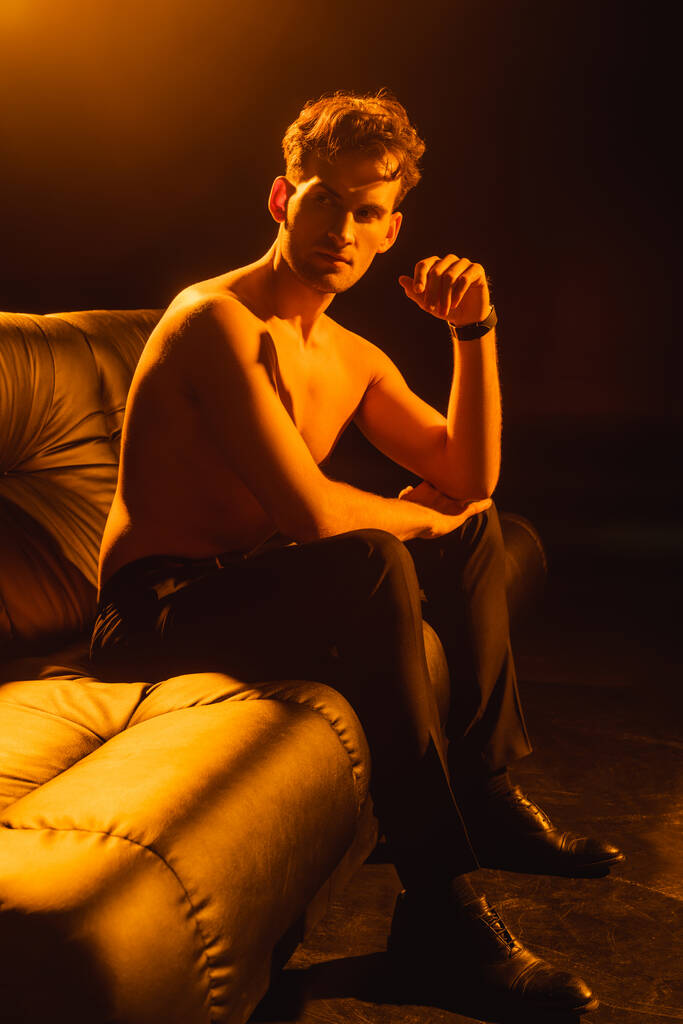 uomo muscoloso guardando lontano mentre seduto sul divano di nero  - Foto, immagini
