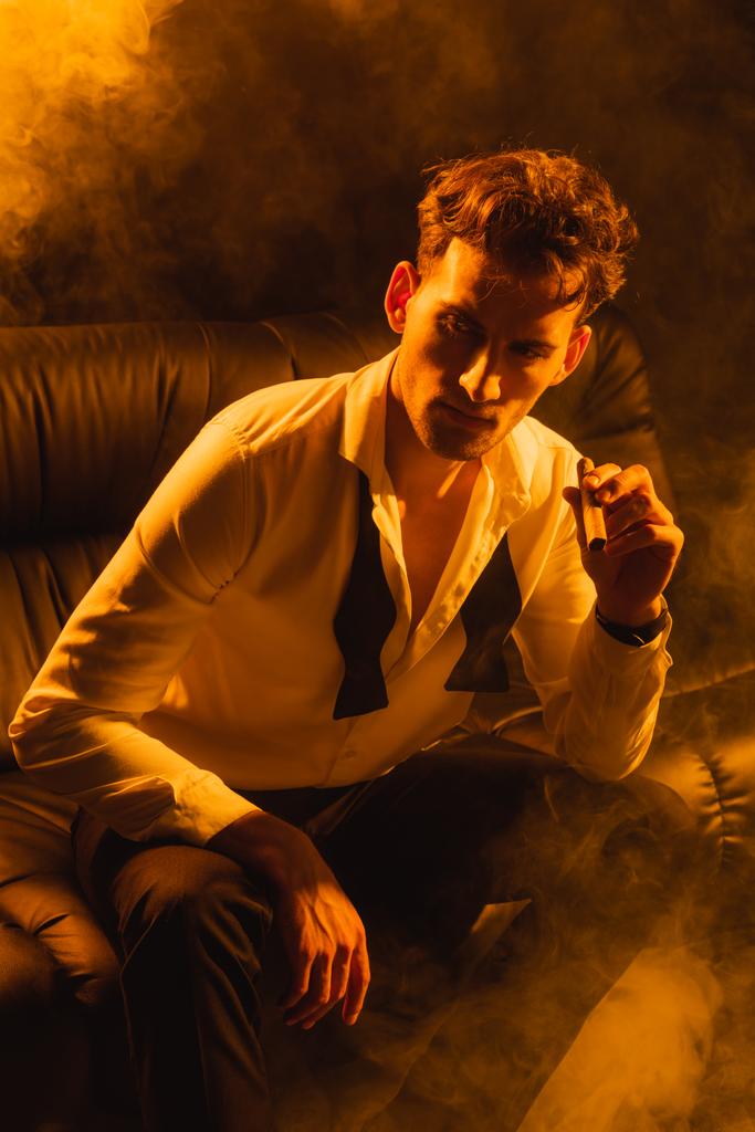 homme en costume assis sur le canapé et tenant cigare sur noir avec de la fumée - Photo, image