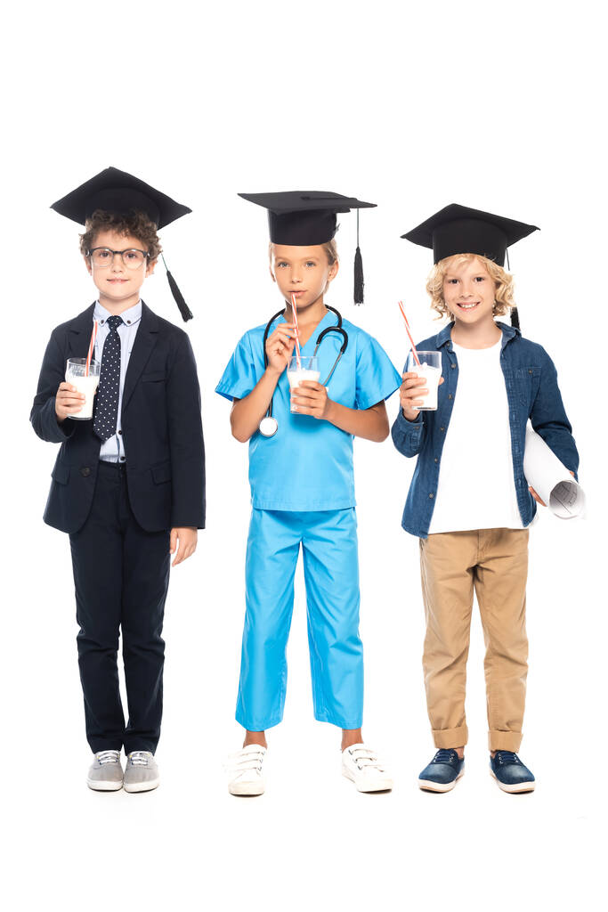 enfants en casquettes de graduation vêtus de costumes de différentes professions tenant des lunettes avec du lait isolé sur blanc  - Photo, image