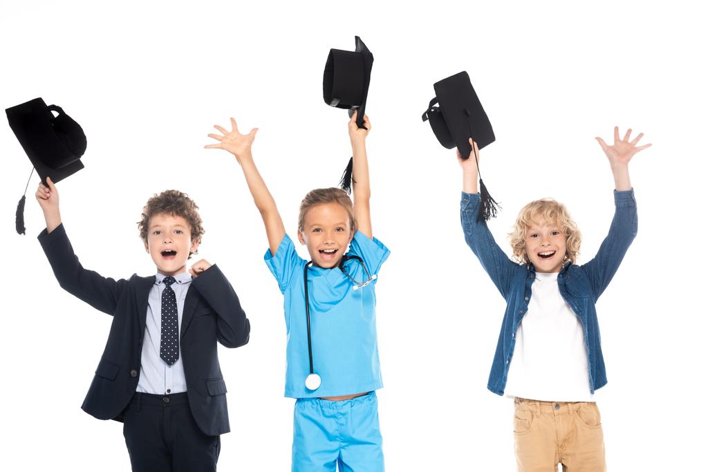 excited kids dressed in costumes of different professions holding black graduation caps above heads isolated on white  - Photo, Image