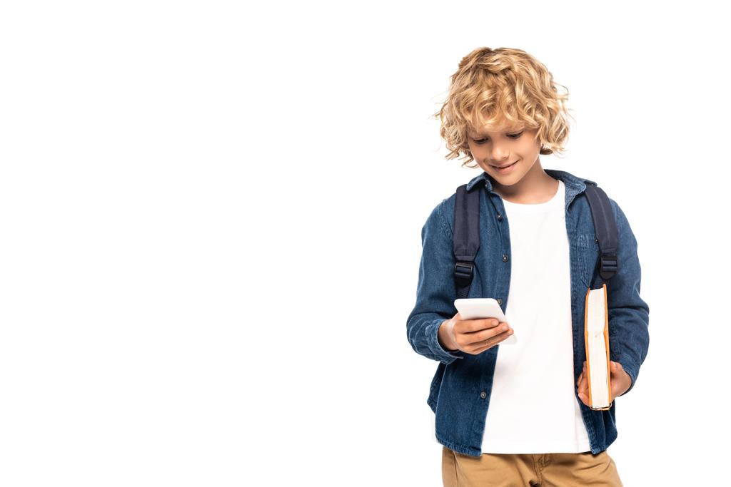 Blonder und lockiger Schuljunge hält Buch in der Hand und benutzt Smartphone isoliert auf weiß  - Foto, Bild