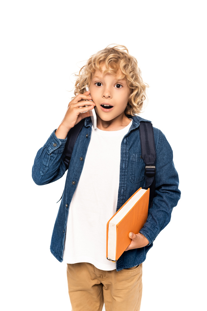 sorprendido y rubio colegial sosteniendo libro y hablando en el teléfono inteligente aislado en blanco  - Foto, imagen