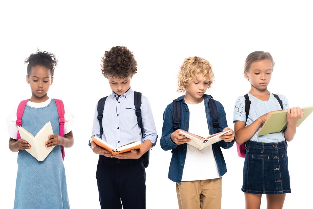 multikulturelle Schulkinder mit Rucksäcken, die Bücher lesen, isoliert auf weiß  - Foto, Bild