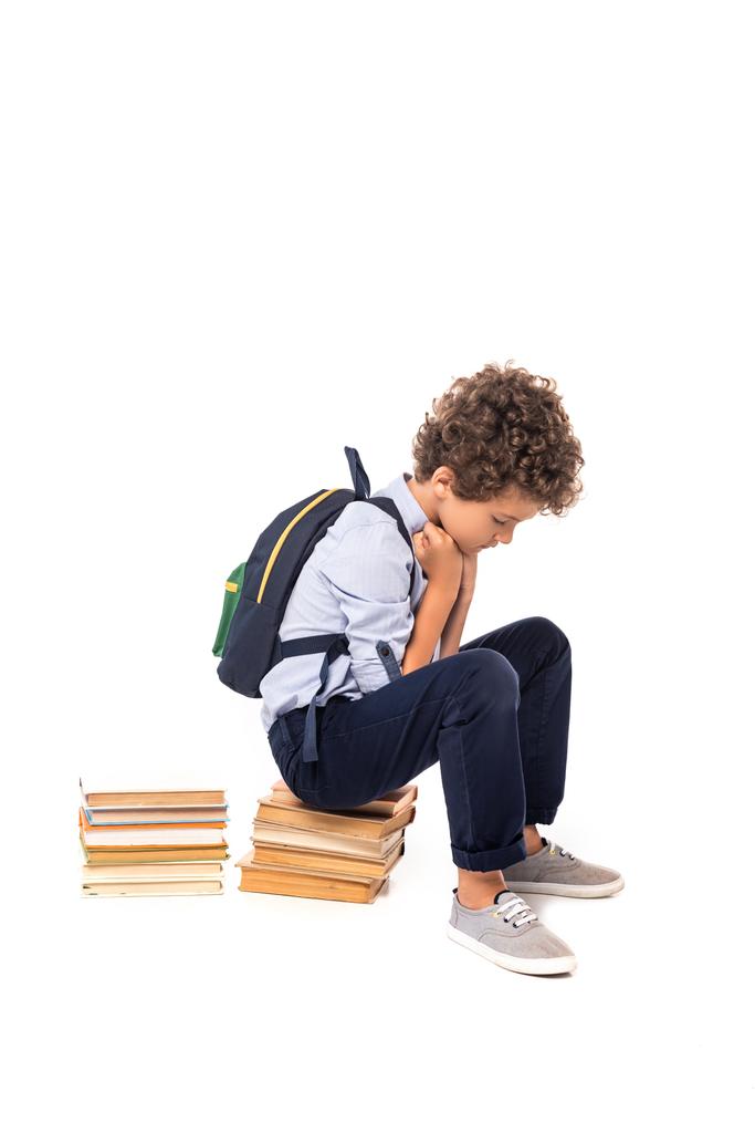 upset schoolboy with backpack sitting on books isolated on white  - Photo, Image