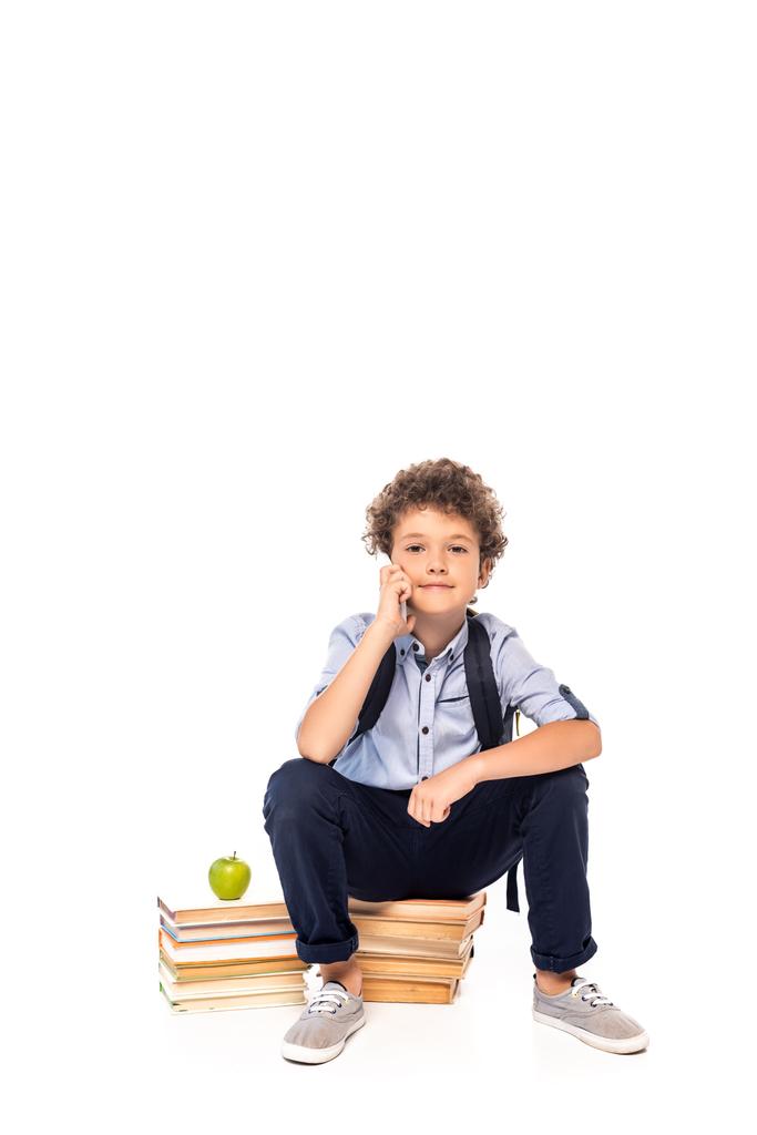 kręcone uczennica z plecakiem siedzi na książkach w pobliżu jabłka i rozmawia na smartfonie odizolowany na białym  - Zdjęcie, obraz