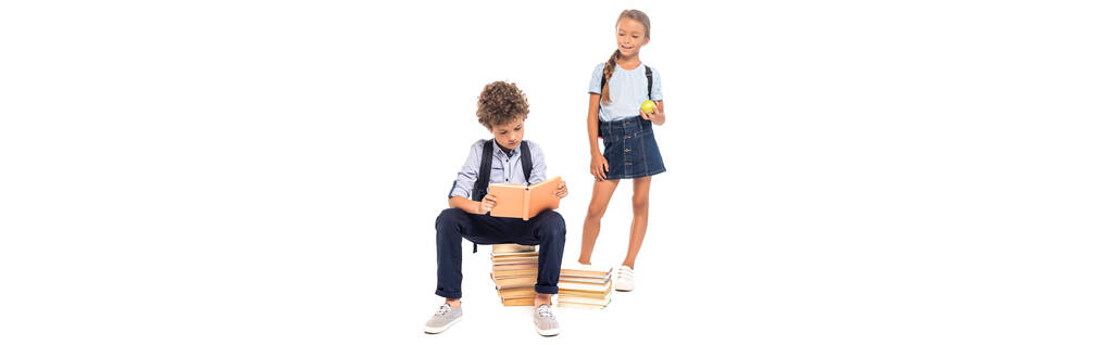 panoramisches Konzept der Schülerin, die einen Apfel in der Hand hält und einen Schüler sieht, der auf Büchern sitzt und isoliert auf weißem Papier liest  - Foto, Bild