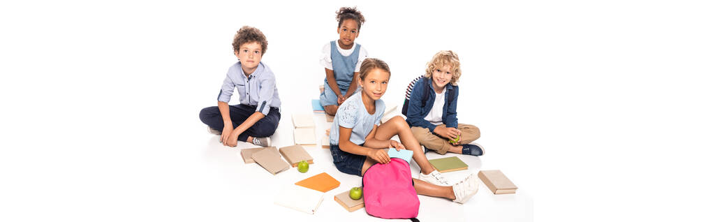 πανοραμική καλλιέργεια των μαθητών που κάθονται κοντά σε βιβλία, μήλα και αφρικανικής Αμερικής παιδί σε ασύρματα ακουστικά που απομονώνονται σε λευκό - Φωτογραφία, εικόνα