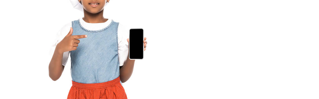 panorama-ernte von afrikanisch-amerikanischen kind in kostüm des kochs zeigt mit finger auf smartphone mit leerem bildschirm isoliert auf weiß  - Foto, Bild