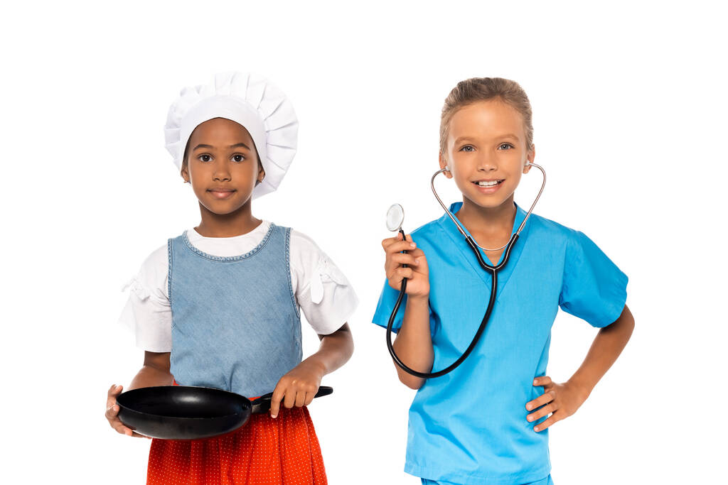 multikulturelle Kinder in Kostümen verschiedener Berufe mit Bratpfanne und Stethoskop isoliert auf weißem Grund  - Foto, Bild