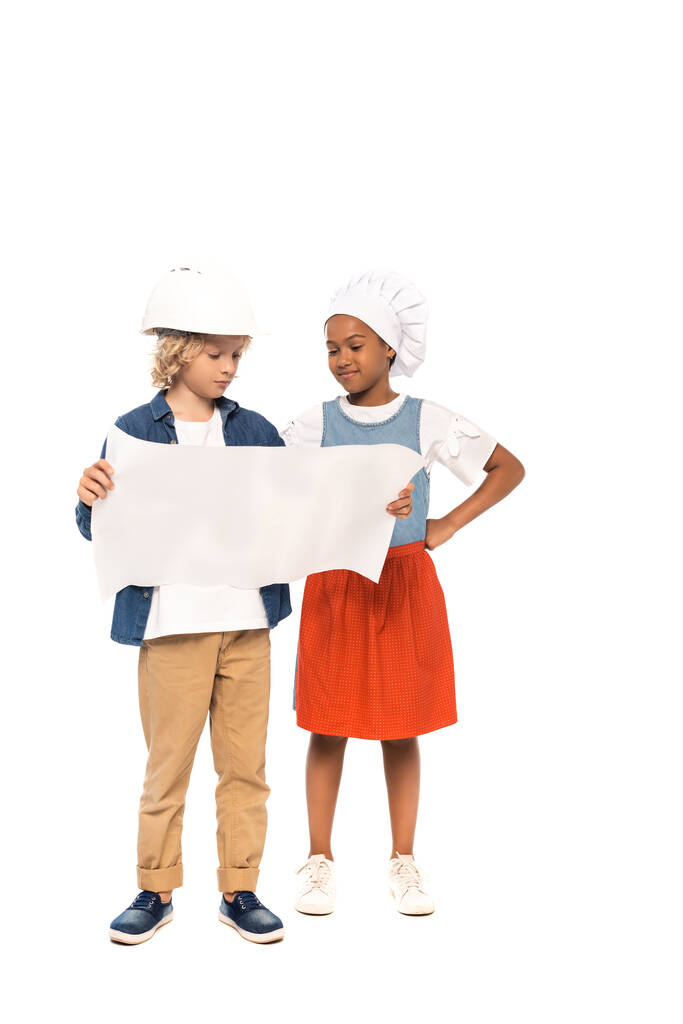 krullende jongen in veiligheid helm en kostuum van de architect kijken naar blauwdruk in de buurt van Afrikaans Amerikaans kind in chef-kok hoed staande met de hand op heup geïsoleerd op wit  - Foto, afbeelding