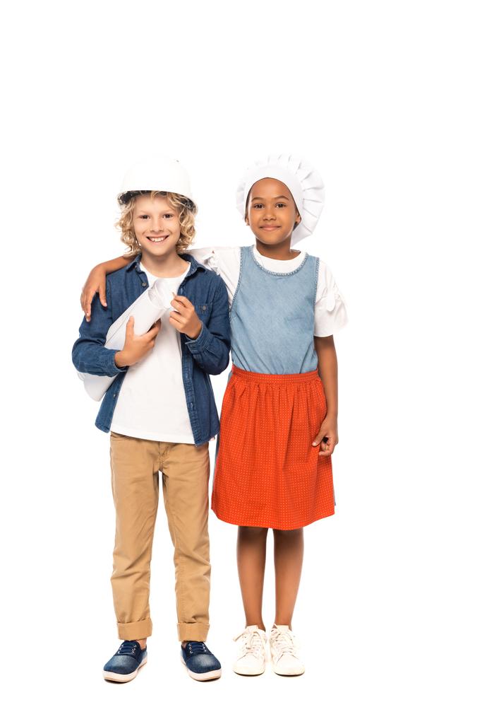 criança afro-americana em chapéu de chef abraçando menino encaracolado em capacete de segurança e traje de arquiteto de pé com planta isolada em branco  - Foto, Imagem