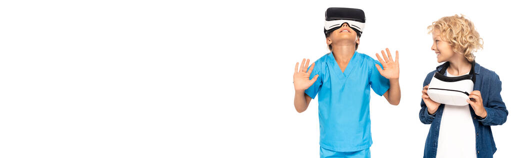 panoramisch concept van krullende jongen kijken naar kind in virtual reality headset gebaar geïsoleerd op wit  - Foto, afbeelding