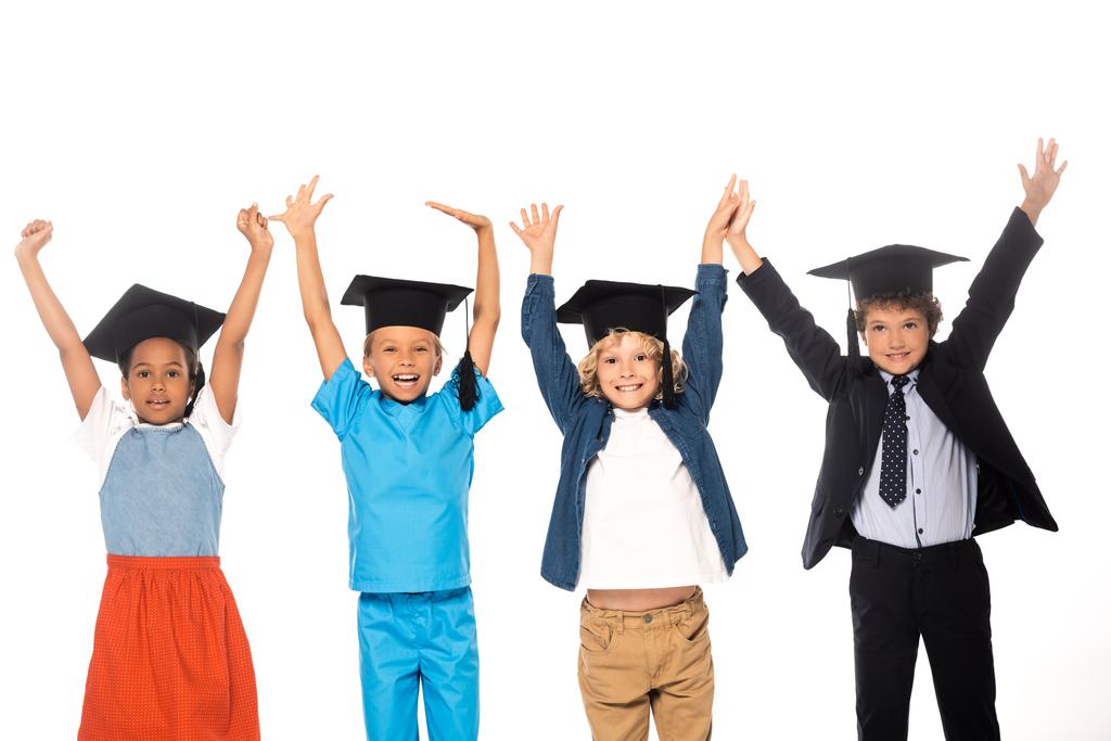 niños multiculturales en gorras de graduación vestidos con disfraces de diferentes profesiones de pie con las manos levantadas aisladas en blanco  - Foto, imagen