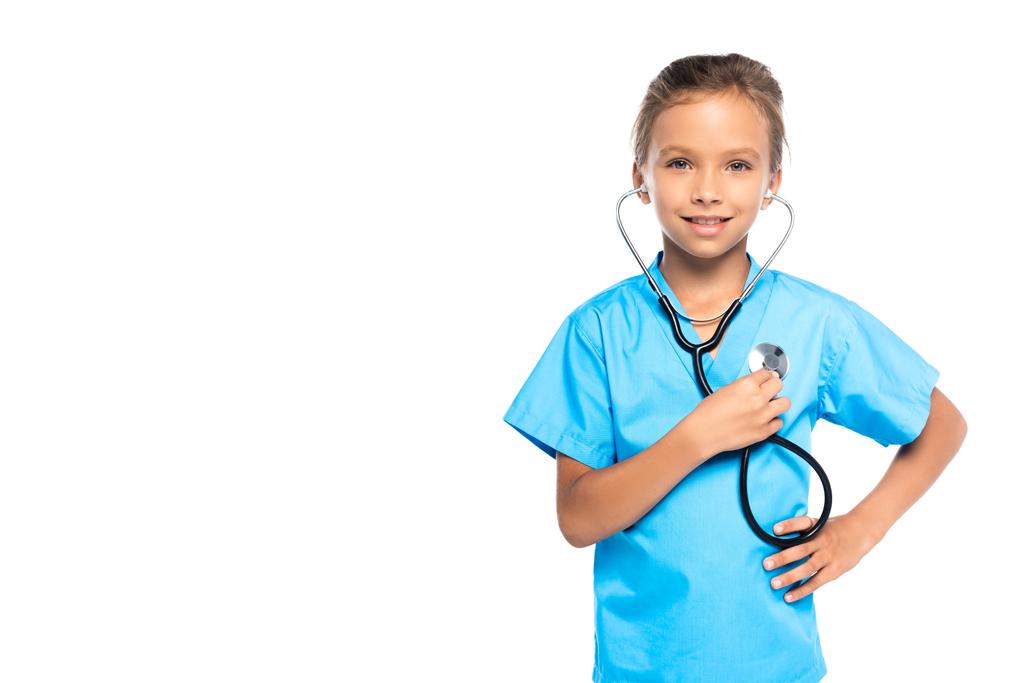 enfant en costume de médecin tenant stéthoscope debout avec la main sur la hanche isolé sur blanc  - Photo, image