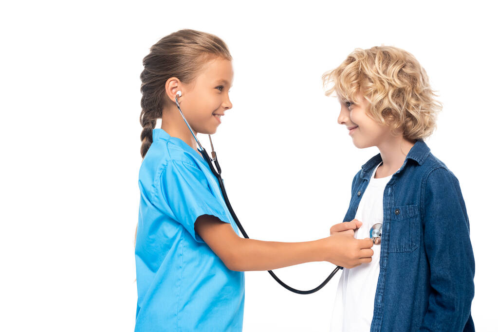 vista lateral de niño en traje de médico examinando chico rubio aislado en blanco  - Foto, imagen