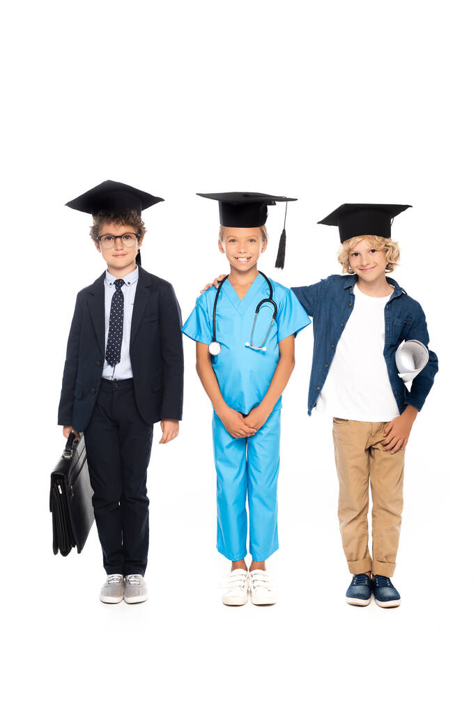 crianças em bonés de formatura vestidas com trajes de diferentes profissões em pé com estetoscópio, planta e maleta isoladas em branco  - Foto, Imagem