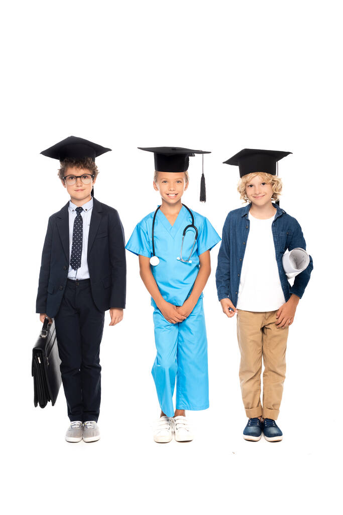 παιδιά σε καπέλα αποφοίτησης ντυμένα με κοστούμια διαφόρων επαγγελμάτων που στέκονται με στηθοσκόπιο, σχέδιο και χαρτοφύλακα απομονωμένα σε λευκό  - Φωτογραφία, εικόνα