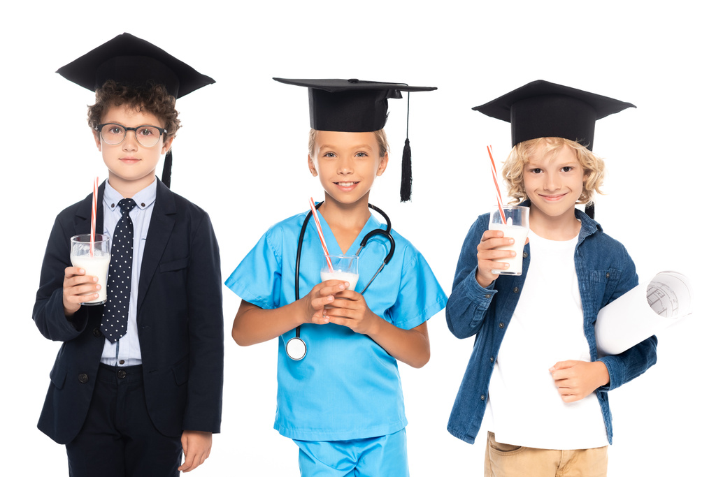 niños en gorras de graduación vestidos con disfraces de diferentes profesiones sosteniendo vasos con leche fresca aislada en blanco  - Foto, Imagen