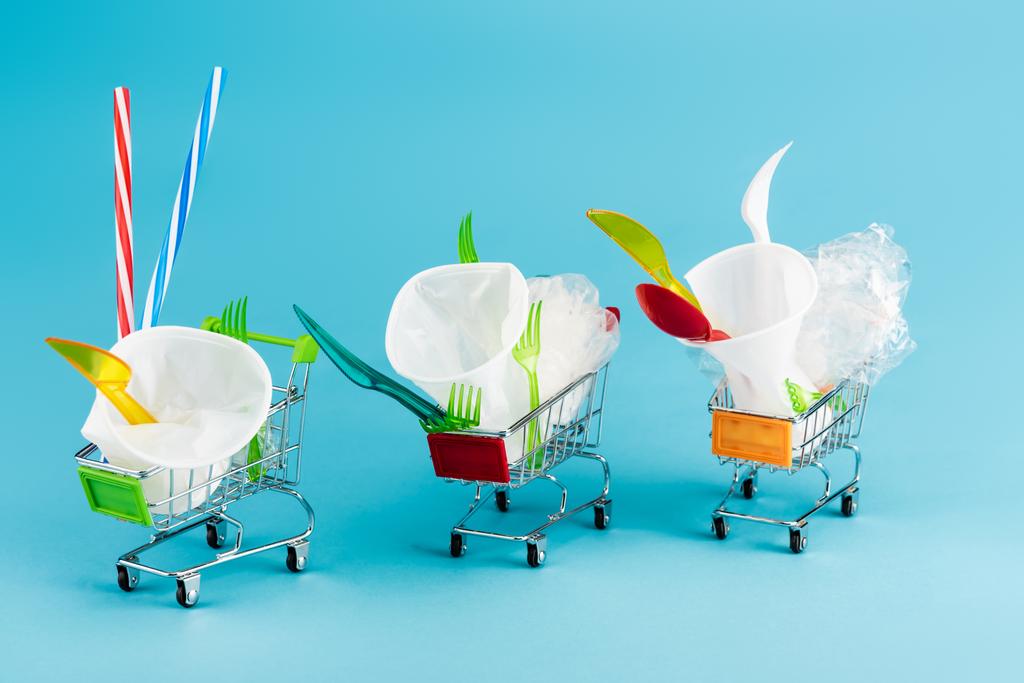 objetos de plástico desechables en pequeños carritos de compras sobre fondo azul - Foto, imagen