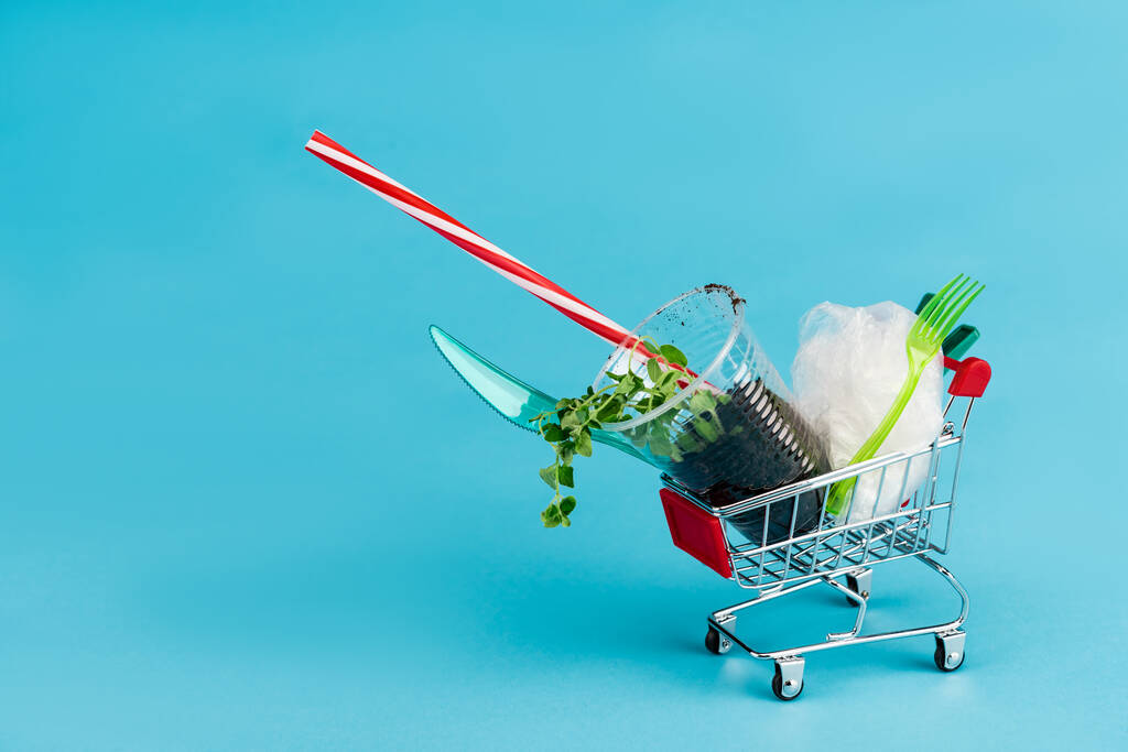 disposable plastic objects and plant in ground in small shopping cart on blue background - Photo, Image