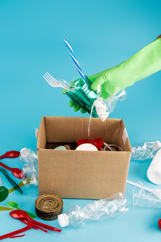 Lastik eldivenli temizleyicinin mavi arkaplandaki karton kutuda çöp toplaması. - Fotoğraf, Görsel