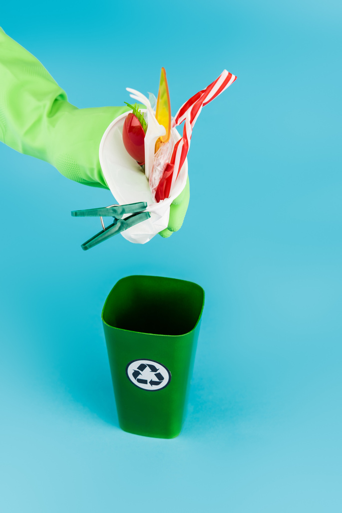 обрезанный вид чистящего средства в резиновой перчатке бросая пластиковый мусор в мусорное ведро на голубом фоне, панорамный снимок - Фото, изображение