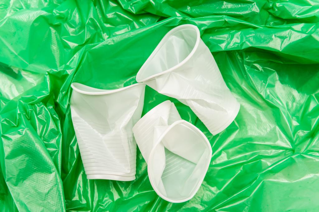bovenaanzicht van witte verkreukelde wegwerpbekers op groene plastic textuur - Foto, afbeelding