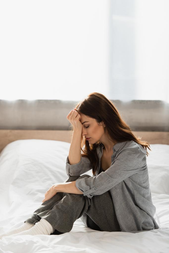 depresja brunetka kobieta z zamkniętymi oczami siedzi na łóżku i dotyka twarzy w domu - Zdjęcie, obraz