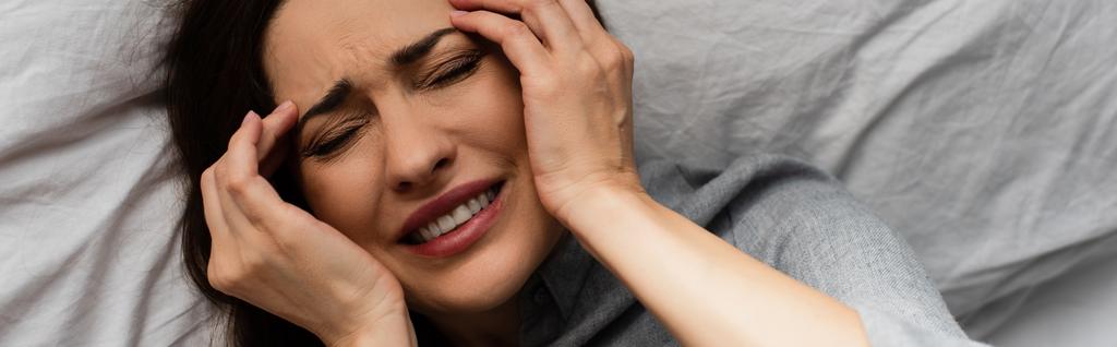 Panoramaaufnahme einer brünetten Frau, die unter Schmerzen leidet und den Kopf berührt, während sie im Bett liegt  - Foto, Bild