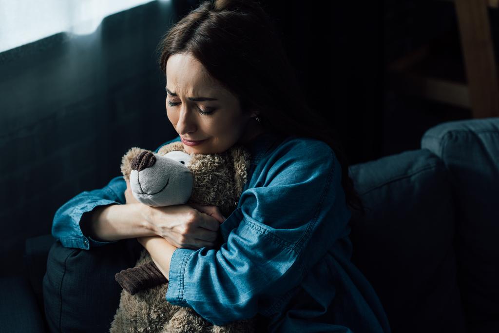 traurige brünette Frau mit geschlossenen Augen hält Teddybär im Wohnzimmer  - Foto, Bild