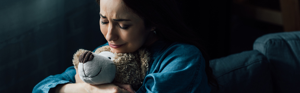 panoramische Ernte von traurigen brünetten Frau mit geschlossenen Augen hält Teddybär im Wohnzimmer  - Foto, Bild