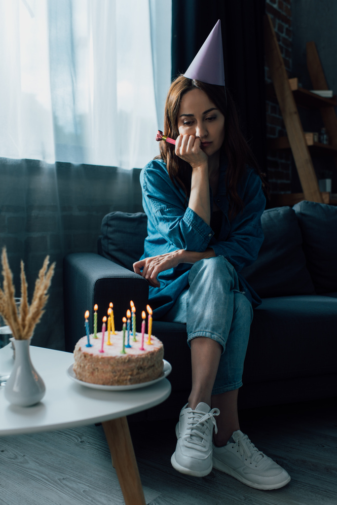 λυπημένη γυναίκα κρατώντας κέρατο κόμμα, κάθεται στον καναπέ και κοιτάζοντας τούρτα γενεθλίων στο τραπέζι του καφέ - Φωτογραφία, εικόνα