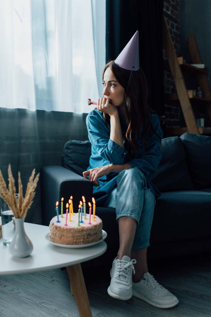 Traurige Frau hält Partyhorn in der Hand und sitzt auf Sofa neben Geburtstagstorte auf Kaffeetisch - Foto, Bild