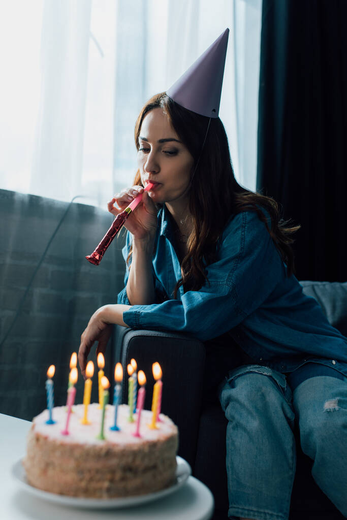 悲しい女性がパーティーホーンを吹いてコーヒーテーブルの誕生日ケーキの近くのソファに座って - 写真・画像