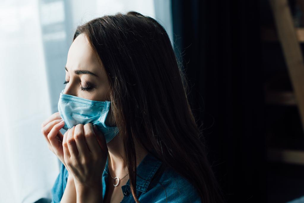 brunette woman with closed eyes touching medical mask at home - Photo, Image