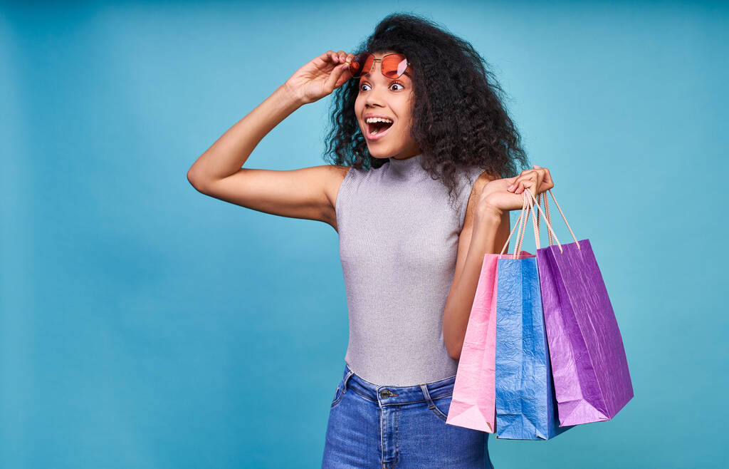 Verkaufskonzept. Aufgeregtes Afro-Mädchen mit roter Sonnenbrille, vielen bunten Einkaufstüten, seitlich mit offenem Mund, isoliert auf hellblauem Hintergrund. - Foto, Bild