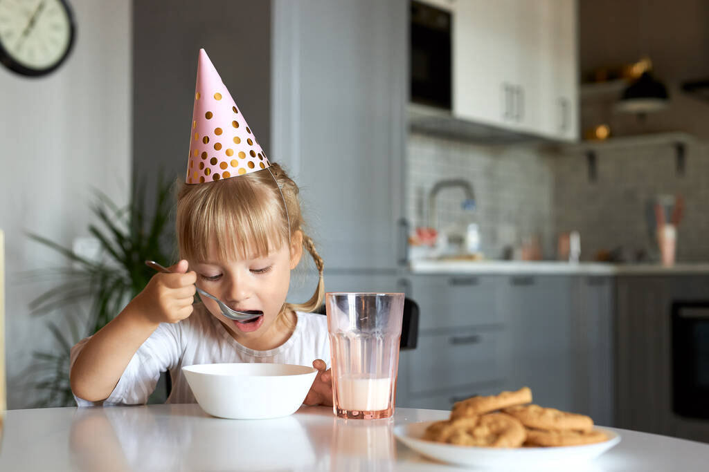 Το κοριτσάκι κάθεται και τρώει πρωινό φορώντας καπέλο για πάρτι. - Φωτογραφία, εικόνα