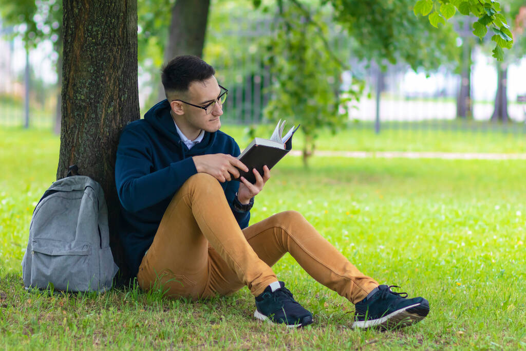 Joven chico concentrado en gafas, estudiante leyendo un libro interesante, apoyado en un árbol, relajándose en el parque de verano con mochila. Guapo inteligente inteligente hombre estudiando al aire libre.  - Foto, imagen